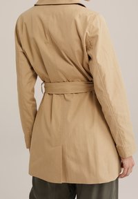 Beżowy trencz z paskiem w talii, długimi rękawami i klasycznym kołnierzem. Tkanina wydaje się gładka, o lekko dopasowanym kroju.