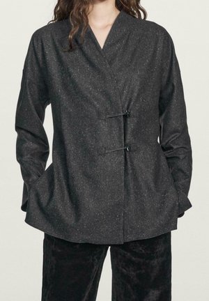 Femme portant une veste portefeuille gris foncé tachetée avec deux fermetures à bascule noires, manches longues et mains dans les poches latérales.