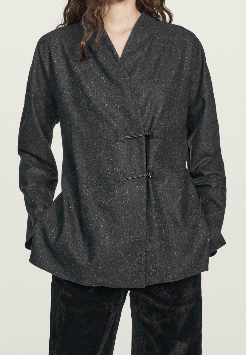 Femme portant une veste portefeuille gris foncé tachetée avec deux fermetures à bascule noires, manches longues et mains dans les poches latérales.