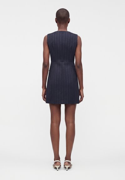 Femme debout de dos, portant une robe sans manches bleu marine à fines rayures et des sandales blanches à petit talon sur un fond uni.