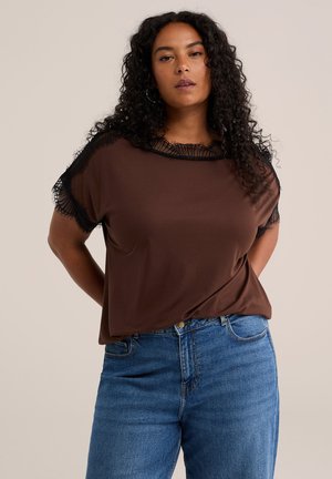 Femme aux cheveux bouclés portant un haut marron à manches courtes avec un bord en dentelle noire et un jean bleu, debout les mains dans le dos.