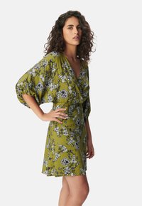 Vestido floral en verde oliva con escote en V y mangas cortas abullonadas. Presenta un estampado floral en blanco y negro y una cintura ajustada.