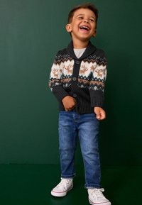 Cardigan na gombíky v sivej farbe s fair isle vzorom, na ktorom sú soby a snehové vločky, skombinovaný s modrými džínami a bielymi teniskami.