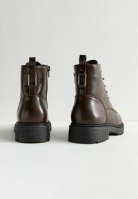Bottines en cuir marron avec une finition lisse, à lacets, et un talon bloc. Présentent une fermeture éclair sur le côté et une sangle décorative avec une boucle.