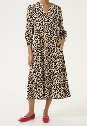 Femme portant une longue robe à imprimé léopard avec des manches 3/4 et des ballerines rouges, debout avec une main dans la poche de la robe.