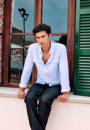 Jaunas vyras su garbanotais plaukais, dėvintis baltą marškinius ir tamsias kelnes, sėdi ant lango palangės su medine rėmu ir žaliais žaliuzėmis lauke.