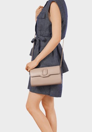 Donna che indossa un vestito azzurro senza maniche con cintura, che porta una borsa a tracolla beige con catena dorata e chiusura ovale.