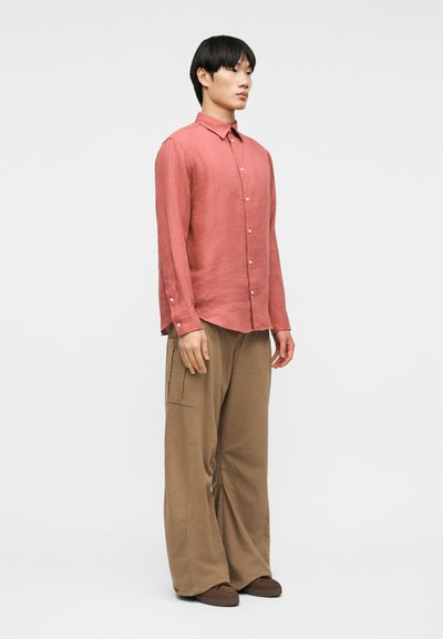 Νέος άνδρας που στέκεται πλάγια, φορώντας ένα μακρυμάνικο κοραλί πουκάμισο με κουμπιά, φαρδιά μπεζ παντελόνια και καφέ παπούτσια σε απλό φόντο.