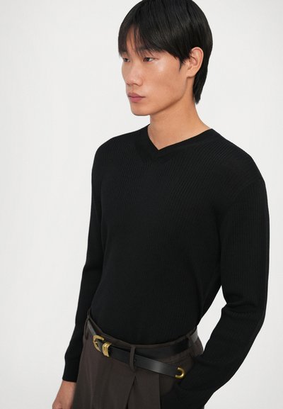 Sweater de malha canelada preta com decote em V, apresentando mangas longas. Usado com calças castanhas e um cinto preto com detalhes em dourado.