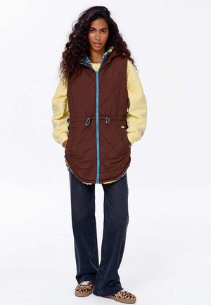 Brązowa pikowana kamizelka z niebieskim zamkiem i ściągaczem w talii, noszona na jasnożółtej bluzie. Ciemne dżinsy i wzorzyste buty dopełniają stylizację.