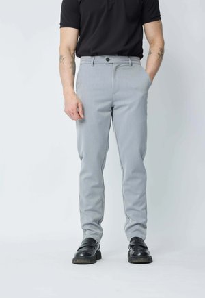 Homme portant un pantalon gris clair, une ceinture noire, un polo noir, des chaussures en cuir noires, se tenant avec une main dans la poche devant un fond uni.