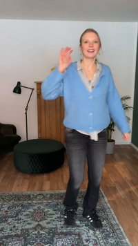 Vrouw met blauwe cardigan en zwarte spijkerbroek zwaait met haar hand binnenshuis nabij een houten kast, een groene poef en een potplant op een patroon tapijt.