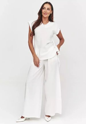 Kobieta z długimi ciemnymi włosami, ubrana w biały krótki top, szerokie białe spodnie i białe szpiczaste obcasy, pozująca z jedną ręką na biodrze.