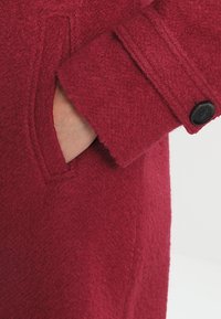 Punainen villakangastakki, jossa on teksturoitu pinta, sivutasku ja napitettu hihansuu. Kangas vaikuttaa lämpimältä ja pehmeältä kosketuksessa.