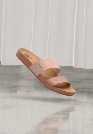 Lyserød dobbeltslynge slidesandal med korkfodsål svævende over en reflekterende grå gulv mod en hvid gardinbaggrund.