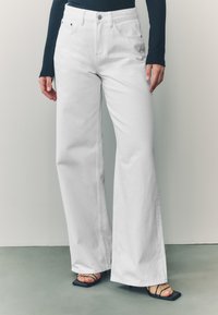Biele džínsy s širokými nohavicami z denimu, s vysokým pásom, päťvreckovým dizajnom a zapínaním na gombíky vpredu. Kombinované s čiernymi sandálmi na opätku so remienkami.