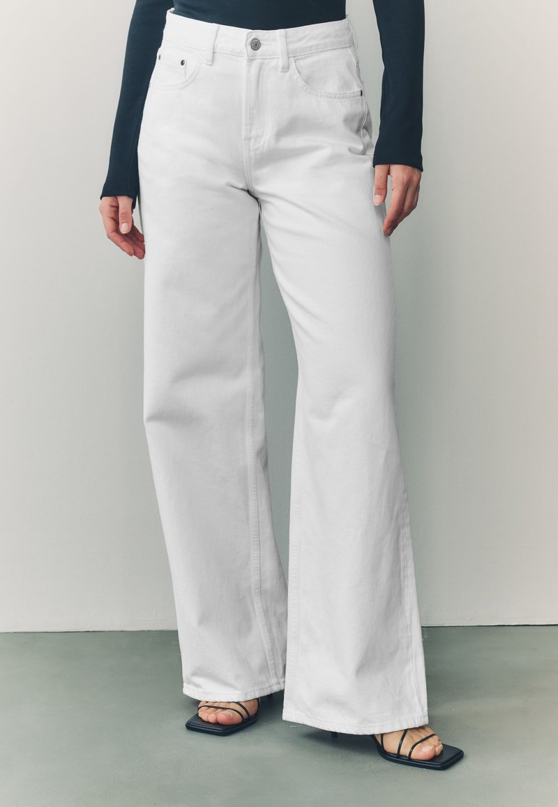 Biele džínsy s širokými nohavicami z denimu, s vysokým pásom, päťvreckovým dizajnom a zapínaním na gombíky vpredu. Kombinované s čiernymi sandálmi na opätku so remienkami.