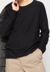 Svart långärmad skjorta i lätt, strukturerad tyg med rund halsringning. Matchad med beige byxor, med en hand vilande i en ficka.