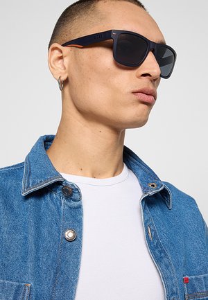 Giovane uomo con occhiali da sole neri, un orecchino a cerchio d'argento, una maglietta bianca e una giacca di jeans blu che guarda verso destra.