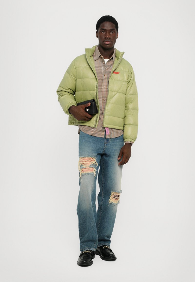 Hellgrüne Pulloverjacke, kariertes Hemd, ausgewaschene blaue Jeans und schwarze Schuhe, mit einer kleinen schwarzen Clutch in einer Hand gehalten.