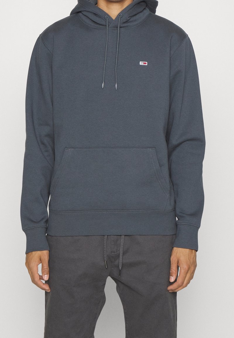 Sudadera gris oscuro hecha de un tejido suave, con un bolsillo frontal tipo canguro, capucha con cordones y un pequeño logo en el pecho. Puños y dobladillo acanalados.