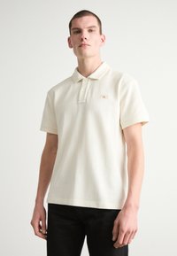 Polo majica kratkih rukava u svijetlo bež pamuku s tri dugmeta i malim CK logom na lijevoj strani prsiju.