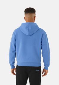 Sudadera azul de textura suave, con capucha ajustable y puños acanalados. Combinada con pantalones negros que tienen un pequeño logo.