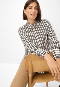 Striped button-up shirt in black and white with long sleeves, paired with beige pants, seated on a light wood chair.