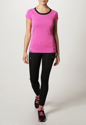 Frau, die geht, trägt ein rosa kurzärmeliges Shirt, schwarze Leggings mit pinken Details und schwarz-pinke Laufschuhe auf hellem Hintergrund.