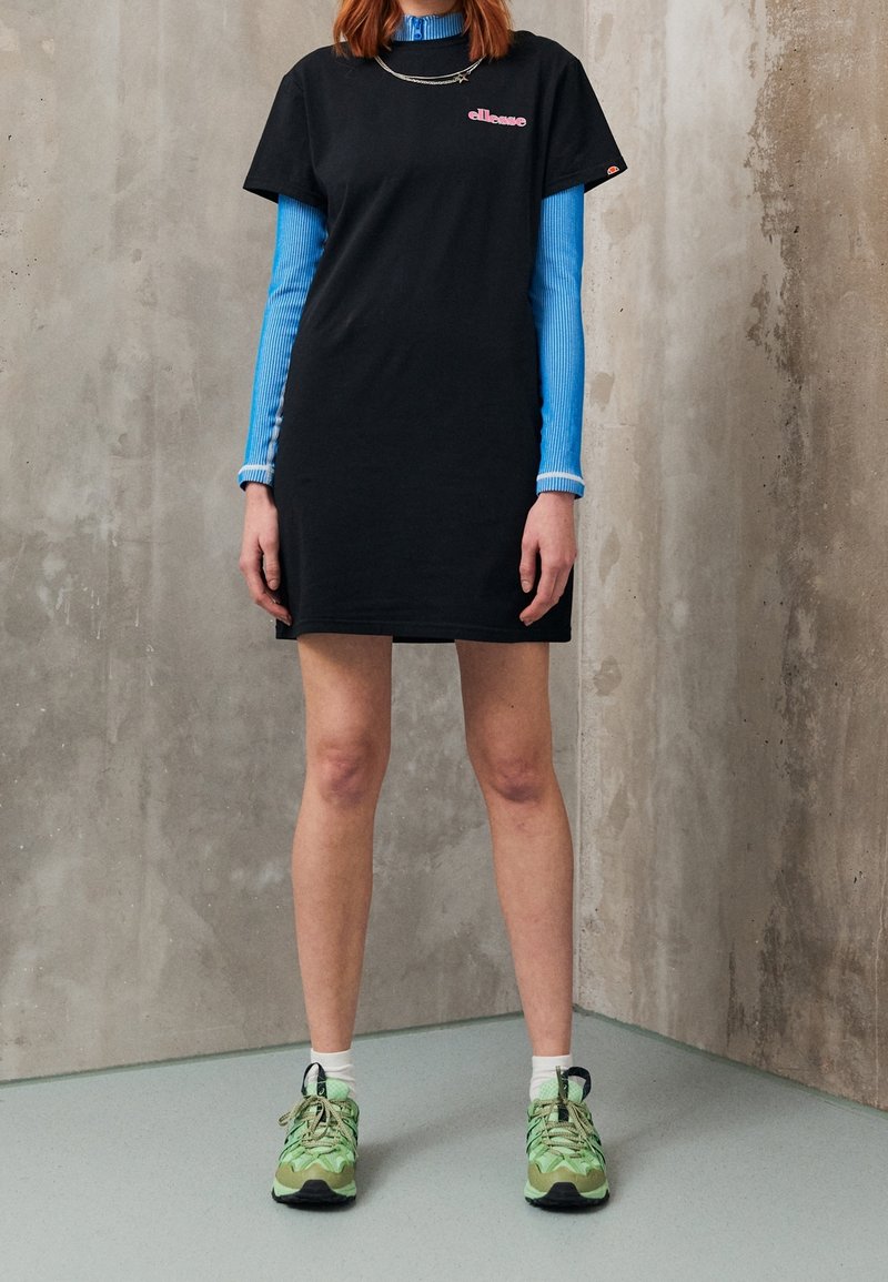 Robe t-shirt en coton noir avec un logo rose, superposée sur un haut côtelé bleu. Portée avec des baskets vertes à motifs et des chaussettes blanches.