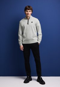 Ljusgrå tröja med hög krage, svart dragkedja och små svarta prickdetaljer, kombinerad med svarta byxor och kraftiga svarta skor.