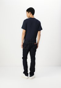 Man wearing a dark navy t-shirt, dark jeans with visible stitching on pockets, and white sneakers standing facing a white wall.