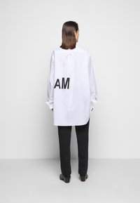 Vit oversized skjorta med en knytning bak och stora svarta bokstäver "AM" på ryggen. Kompletteras med svarta byxor och bruna skor.