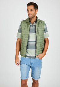 Grüne, gestepptes Weste über einem grauen und grünen gestreiften Poloshirt, kombiniert mit hellblauen, hochgekrempelten Jeansshorts. Einfache Reißverschlussdetails an der Weste.