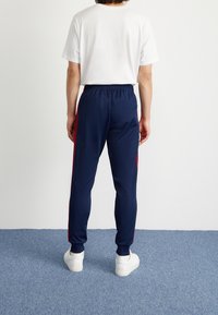 Person som står på en blå matta, klädd i marinblå träningsbyxor med röda sidostreck, vita sneakers och en enkel vit t-shirt, med ryggen mot kameran.