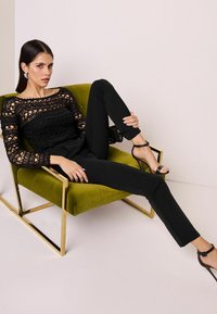 Černý krajkový top se složitými vzory, spárovaný s černými zúženými kalhotami. Model sedí na zelené sametové židli se zlatými kovovými akcenty.