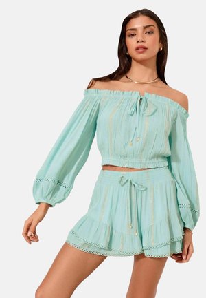 Frau trägt ein hellblaues Off-Shoulder-Top mit langen Ärmeln und passende Shorts mit Rüschen- und Spitzendetails, steht vor einem einfarbigen Hintergrund.