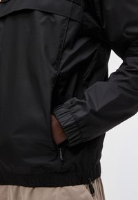 Άτομο που φοράει μαύρο μπουφάν με φερμουάρ και ελαστικά μανίκια, το χέρι μέσα στην τσέπη του μπουφάν, μπεζ παντελόνι ορατό κάτω από το τελείωμα του μπουφάν.