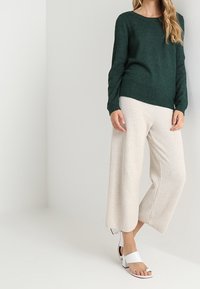 Mörkgrön stickad tröja, krämfärgade vida byxor med texturerat mönster, och vita sandaler med blockklack. Avslappnad passform med rund halsringning.