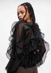 Blusa nera trasparente con ampie decorazioni arricciate lungo le maniche e il retro, tessuto leggero e design fluido.