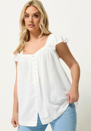 Lyshåret kvinde iført en hvid kortærmet, knappet bluse og lyseblå jeans, stående mod en ensfarvet baggrund.