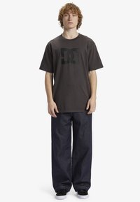 T-shirt gráfica preta com decote redondo, combinada com jeans de denim azul escuro largos. Modelo usa ténis pretos, em pé sobre um fundo branco.