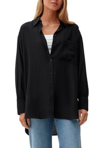 Camicia nera con bottoni, realizzata in tessuto leggero, con colletto, maniche lunghe e taschino sul petto. Abbinata a jeans azzurri chiari.