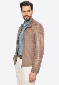 Giacca di pelle marrone con accenti a coste sulle spalle, tasche con zip e design aderente, abbinata a una camicia azzurra con bottoni sotto.