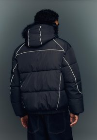 Musta untuvatakki, jossa valkoiset tehosteompeleet, huppu ja tekoturkisreunus. Malli on laatikkomainen ja siinä on resorit hihansuita.