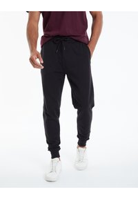 Schwarze Jogginghosen aus weichem Stoff, mit einem Kordelzugbund, Seitentaschen und gerippten Bündchen. Getragen mit weißen Sneakers.