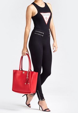Frau trägt ein schwarzes, ärmelloses Guess-Oberteil und enge Hosen mit Reißverzierung, hält eine große rote Handtasche und trägt schwarze Sandalen mit hohen Absätzen.
