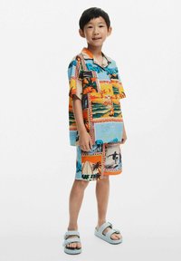 Spalvotas trumpomis rankovėmis marškinėliai ir atitinkantys šortai su paplūdimio ir banglenčių motyvais oranžinės, mėlynos ir geltonos spalvų atspalviuose. Dėvima su šviesiai mėlynais sandalais.