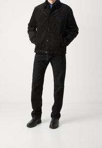 Černá manšestrová bunda s límcem, předním zapínáním na patentky a logem na hrudi. Kombinováno s černými kalhotami a koženými botami.