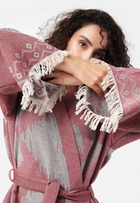 Ružový a šedý tkaný župan s komplexnými geometrickými vzormi, vybavený třásněmi na rukávoch a zodpovedajúcim opaskom, vyrobený z jemnej látky.